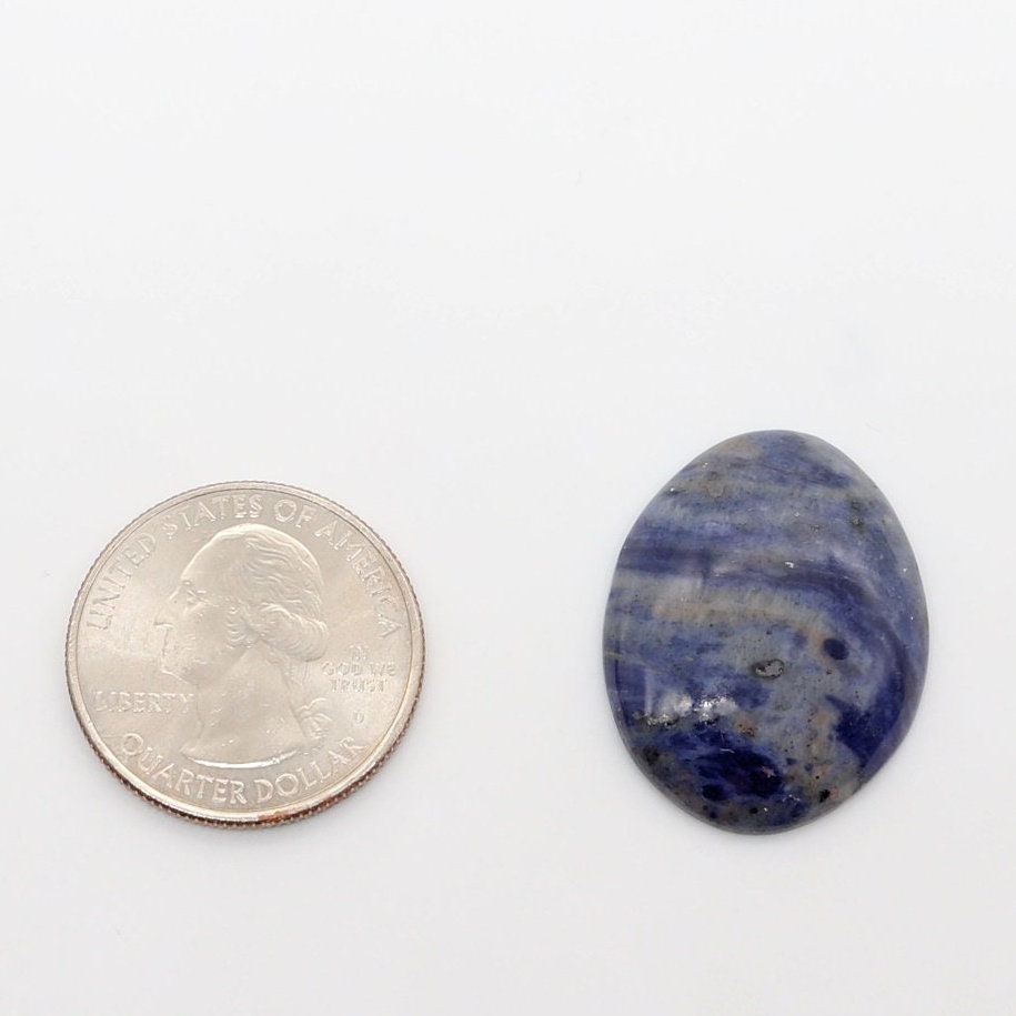 Natural Foidolite cabochon shown beside a U.S. quarter for size comparison, displaying layered deep blue, slate blue and gray natural mineral patterning against a white background.
