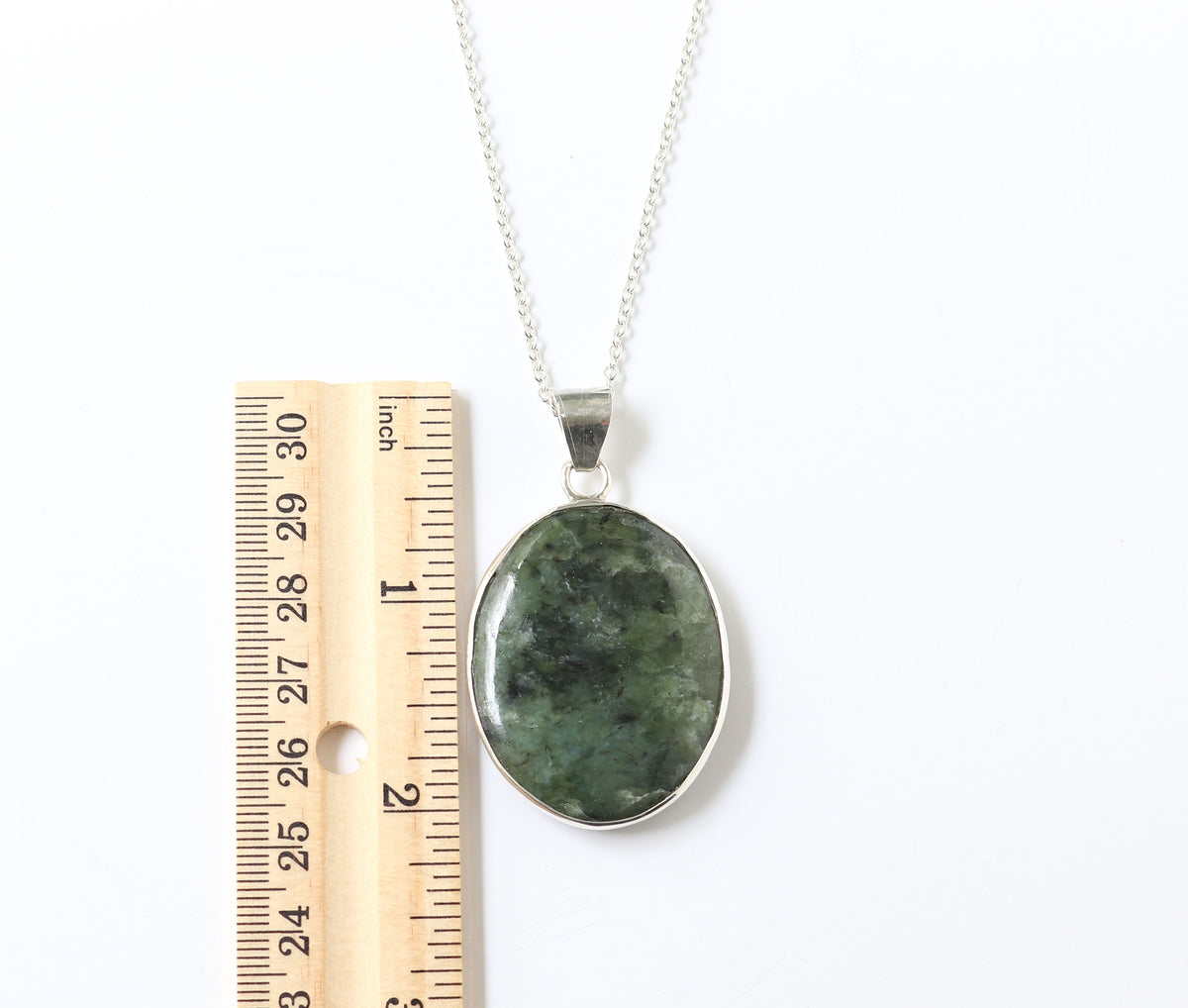 A silver necklace with an oval green stone pendant, shown next to a wooden ruler for scale on a white background.