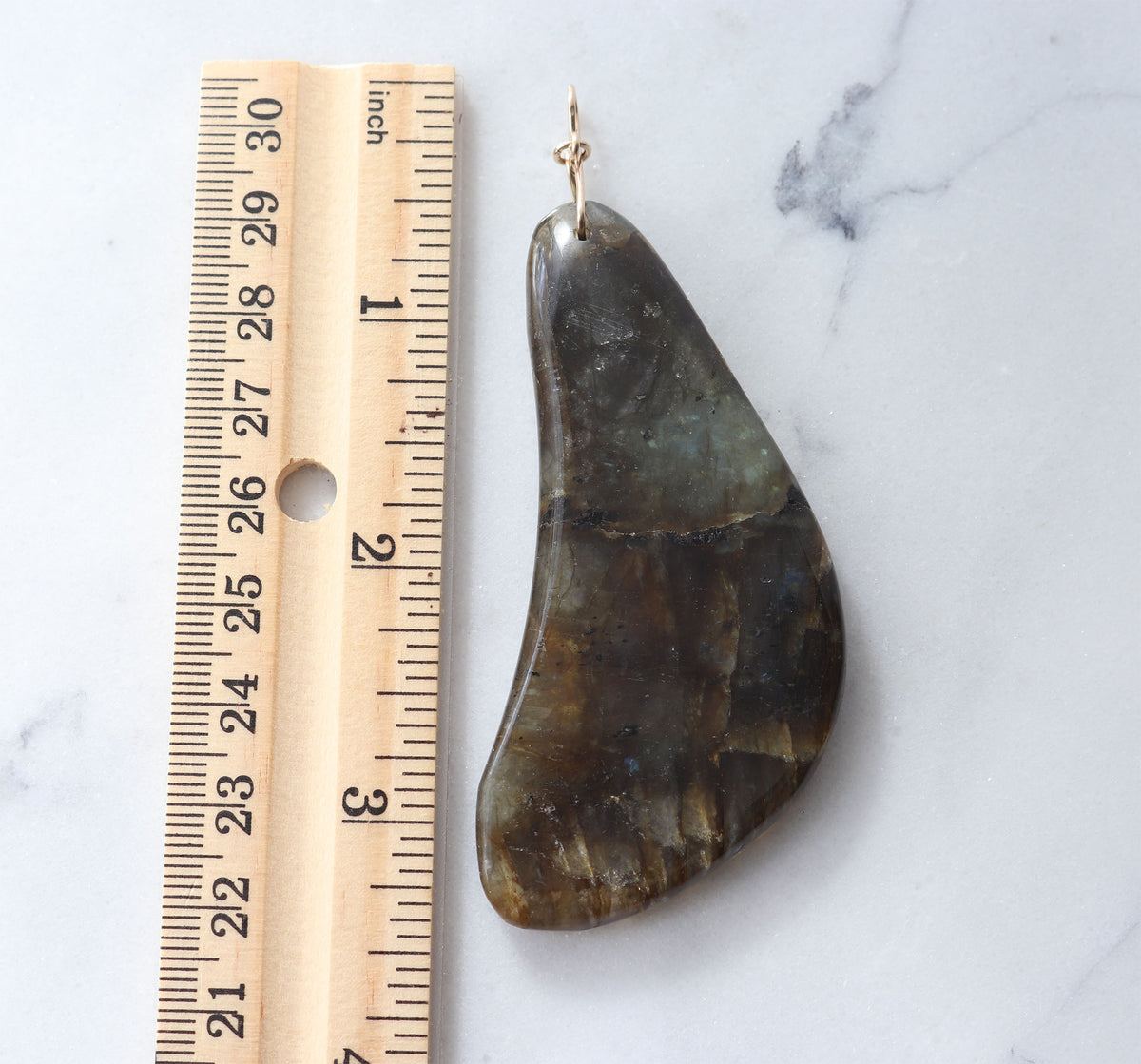A teardrop-shaped polished labradorite gemstone pendant with a gold jump ring, shown next to a wooden ruler on a white marble surface to indicate its size.