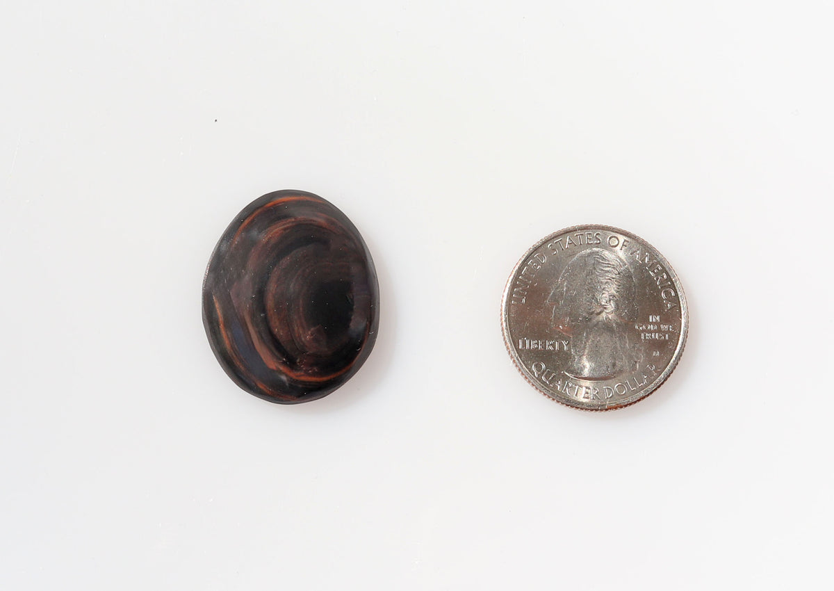 Hand-cut Mahogany Obsidian oval cabochon with reddish-brown and black swirling patterns shown next to U.S. quarter for scale against white background.