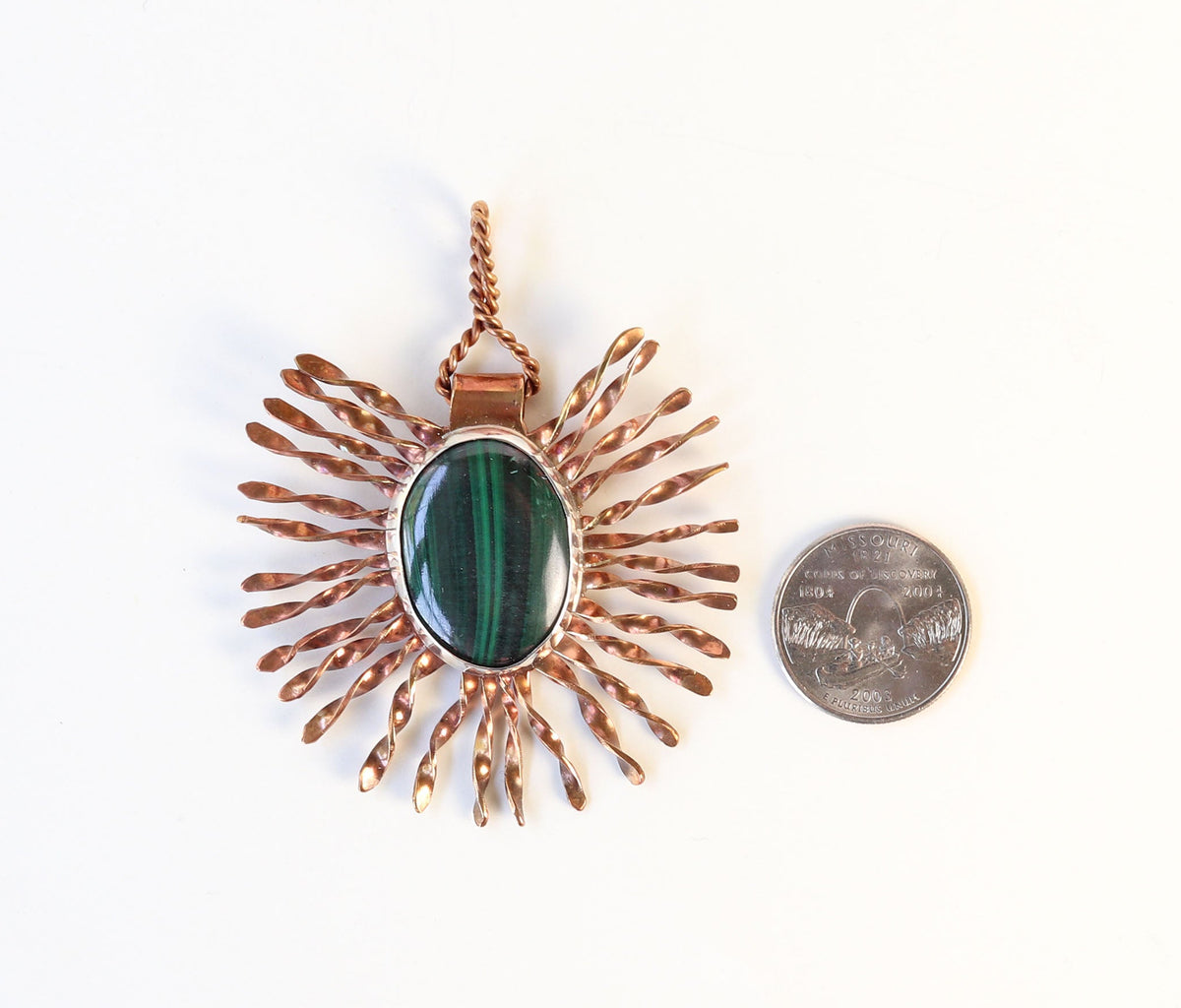 A copper sunburst pendant with a green malachite stone center, shown next to a US quarter coin for scale on a white background.