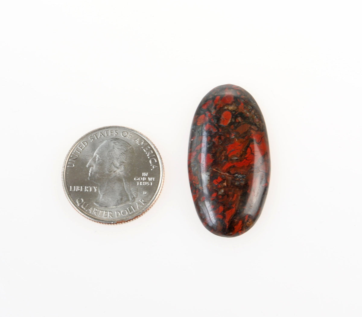 Front view of Natural Red and Black jasper gemstone cabochon, hand-cut and polished into an oval shape to reveal natural mottled patterns of rich reds, earthy blacks and a small white inclusion within the stone.  Shown next to US quarter for scale against a white background.