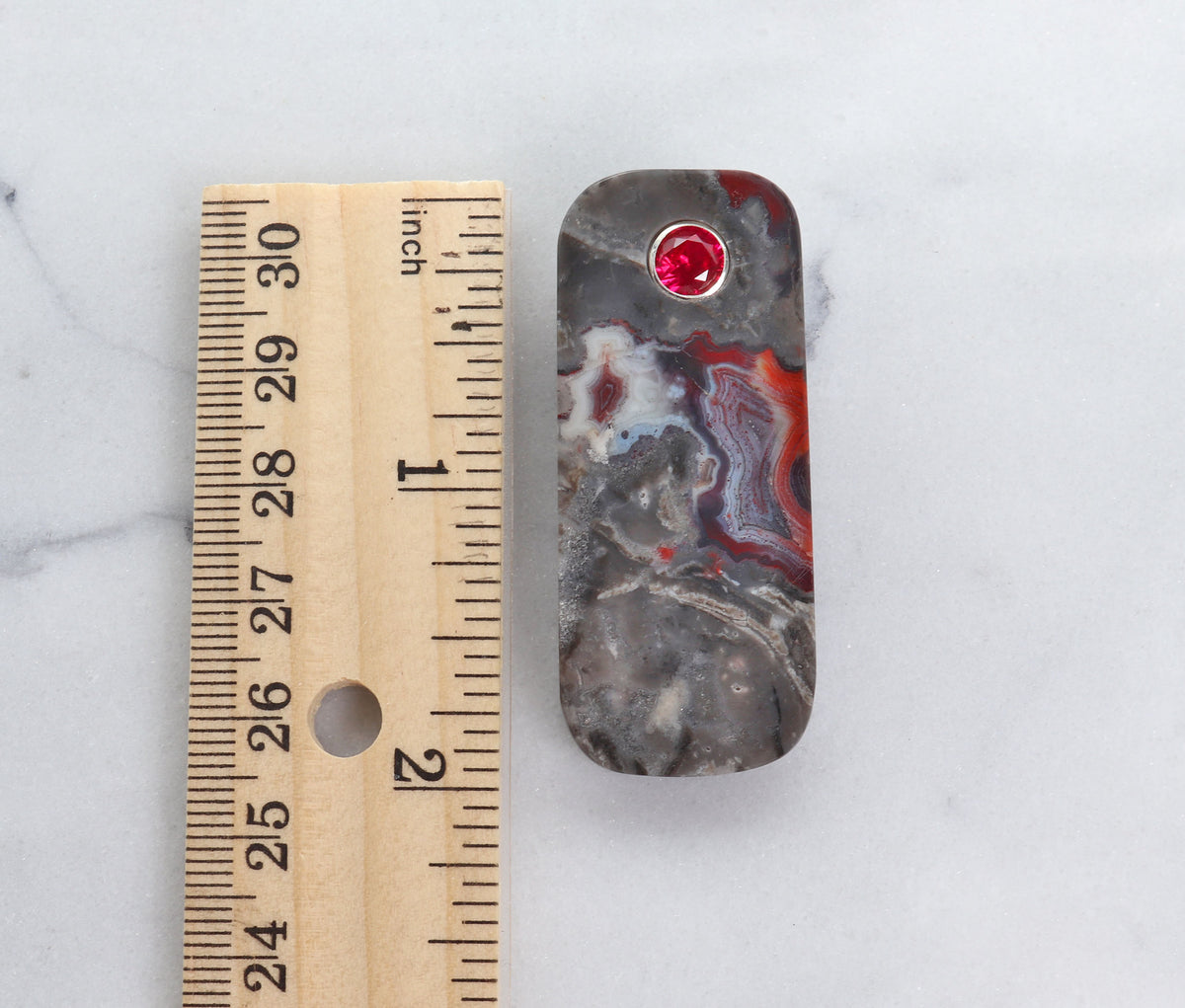 A Red Mexican Agate pendant with 6mm lab-grown ruby inlay and Argentium Silver bail shown next to ruler for size against a white marble slab background.