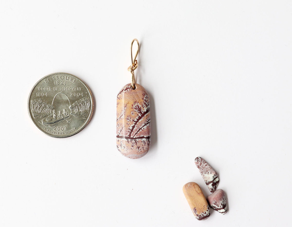 A Sonoran Dendritic Rhyolite pendant with warm tan and soft pink tones laced with delicate white and deep brown dendritic patterns finished with a custom 14/20 gold-filled infinity bow bail.  Shown on white background next a few raw Sonoran Dendritic pebbles for props and Missouri US quarter for scale.
