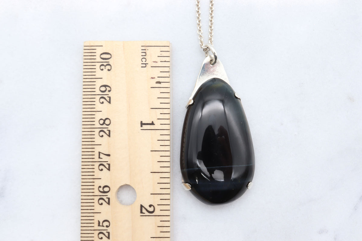 A Handcrafted African Blue Tiger's Eye teardrop pendant with deep blue-black reflective gemstone, solid Argentium 935 silver tabbed setting and silver chain., shown next to a wooden ruler for scale on a white marble background.