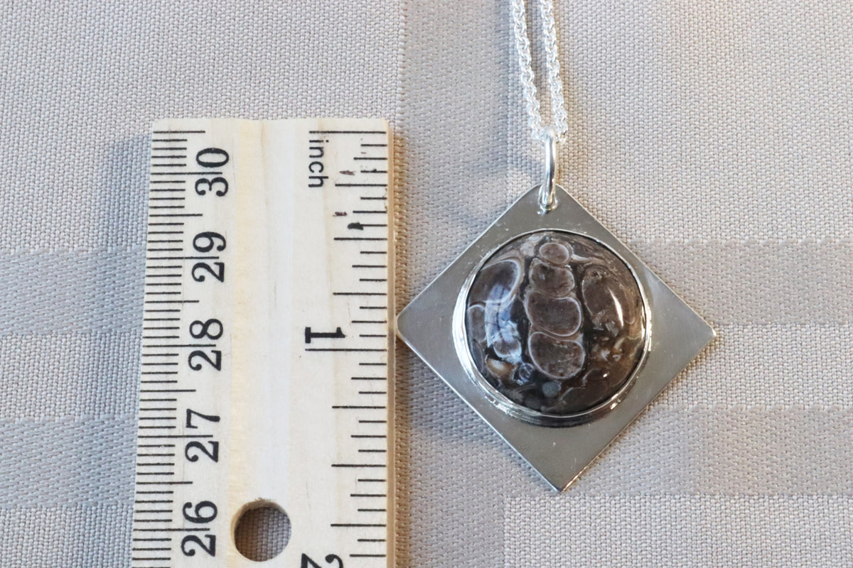 Natural Turritella Agate fossil pendant in Argentium 935 Silver with Argentium silver chain, shown next to a wooden ruler for scale on a light gray textured fabric background.