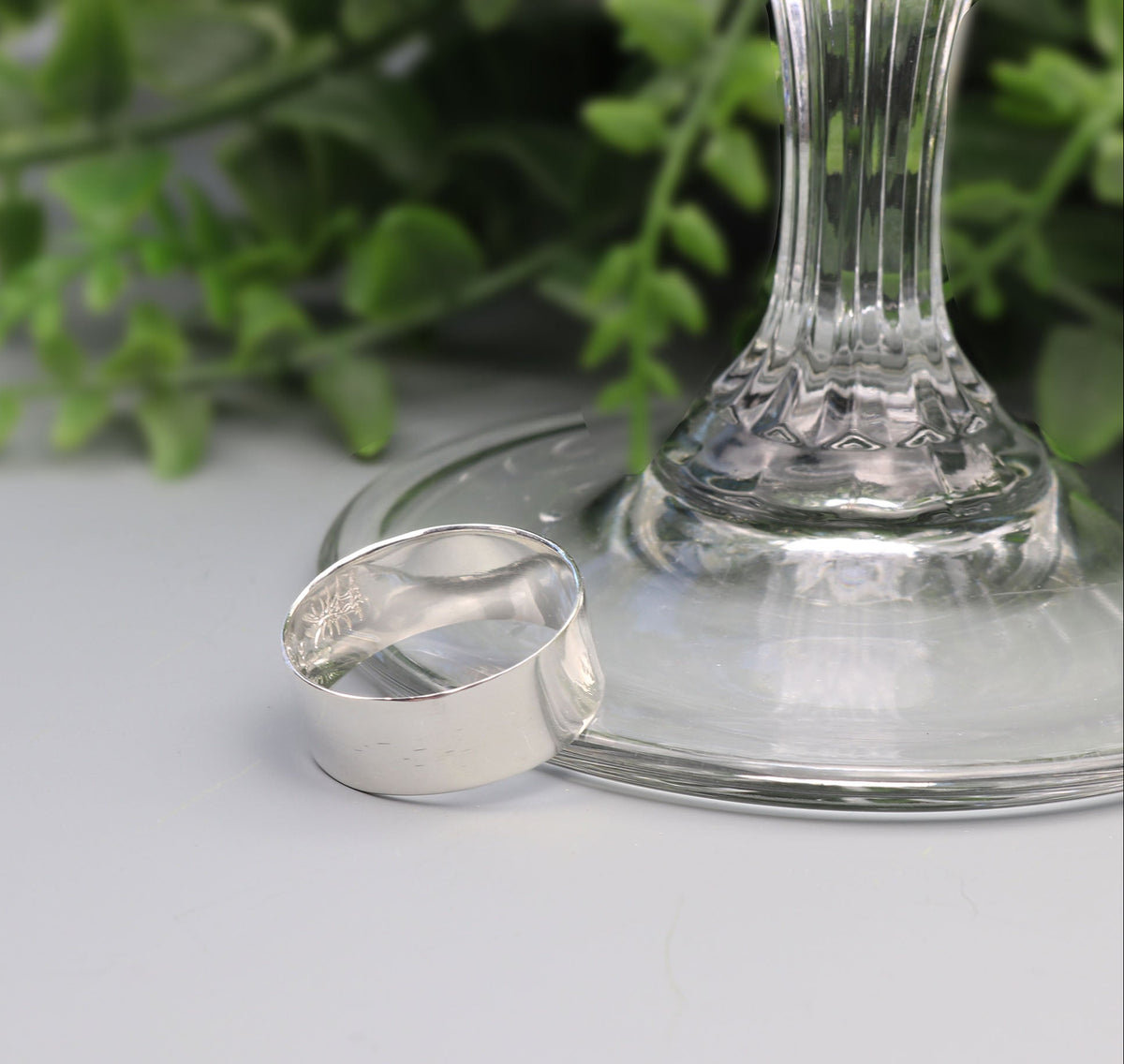 A polished silver ring with a hammered texture, resting on the base of a clear glass stem against a light gray background with blurred green foliage.