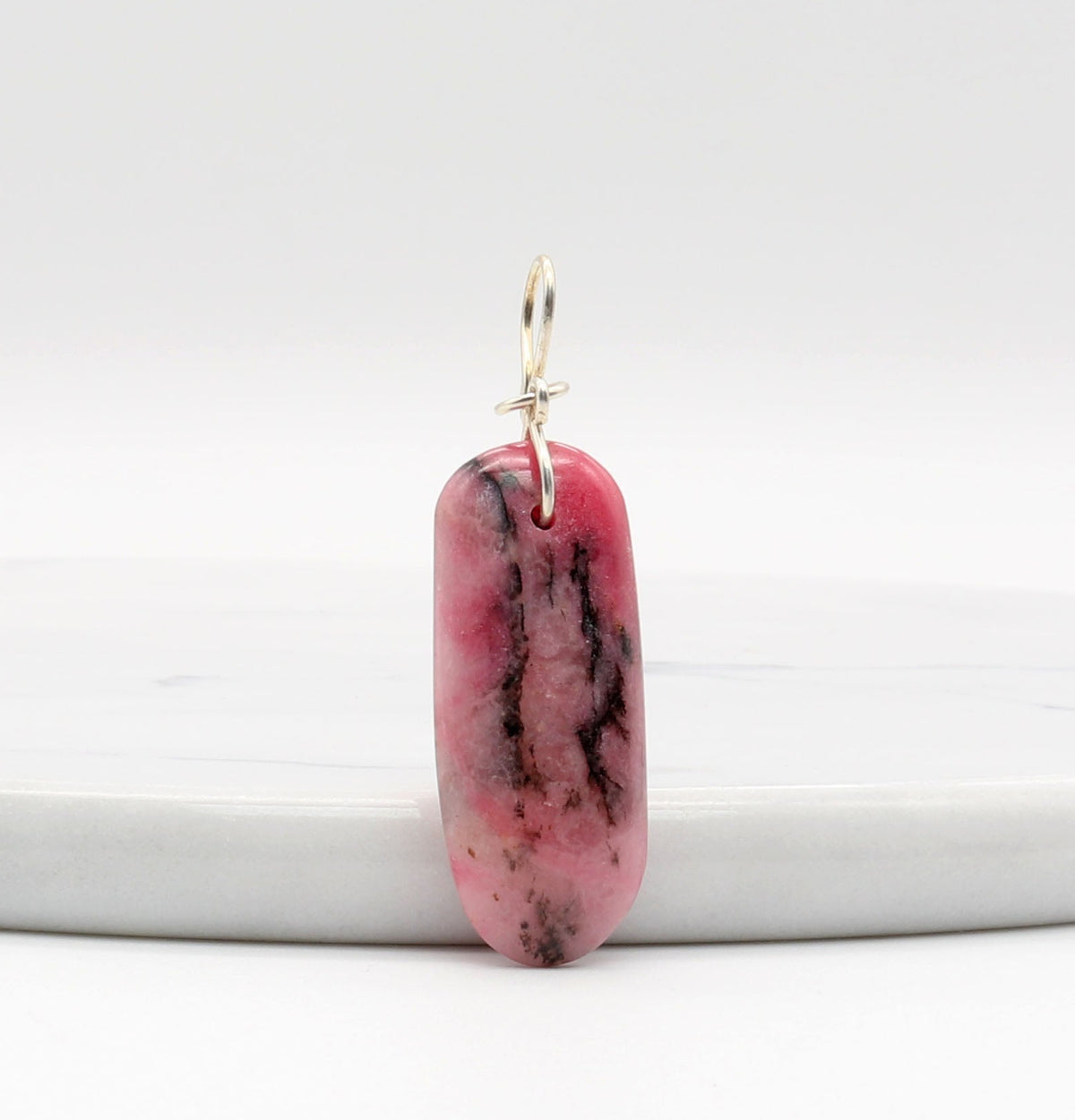 A pink rhodonite gemstone with black inclusions designed with a silver bail. The stone is a long oval shape with a smooth surface.  Shown against a white marble slab.