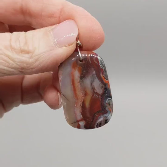 Demo of Red Mexican Agate pendant features of  deep red and black tones with natural flowing banding and a Subtle translucency that catches the light.  Shown on light gray background.