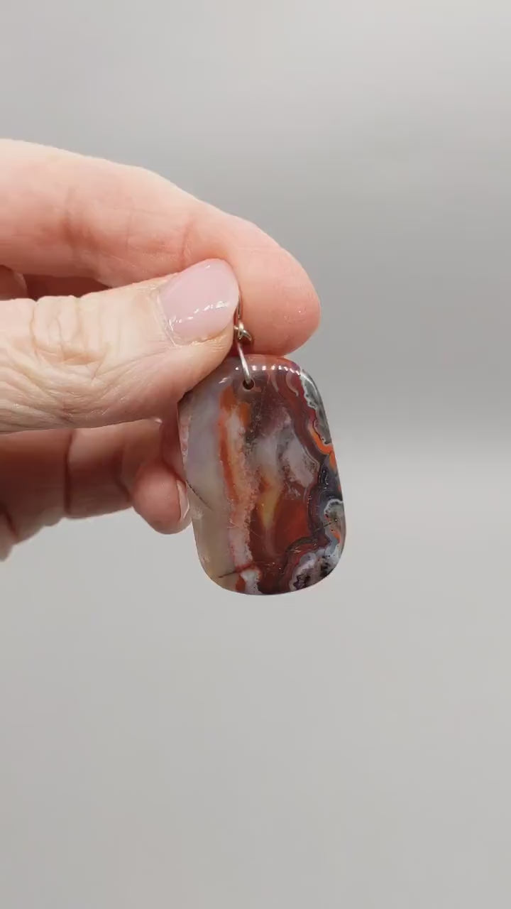 Demo of Red Mexican Agate pendant features of  deep red and black tones with natural flowing banding and a Subtle translucency that catches the light.  Shown on light gray background.