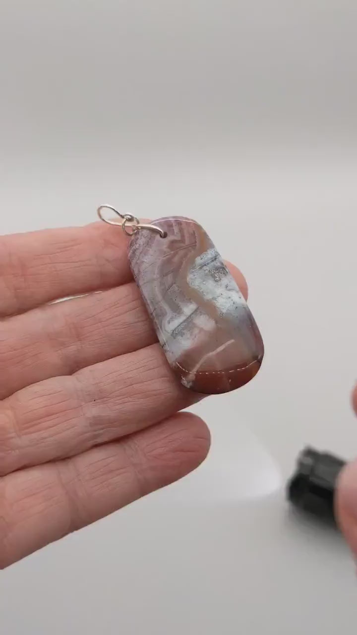 Demo clip of a Red Mexico Agate light display of the translucent features of this mauve and white  tage shaped pendant with Argentium wire loop shown against white background.