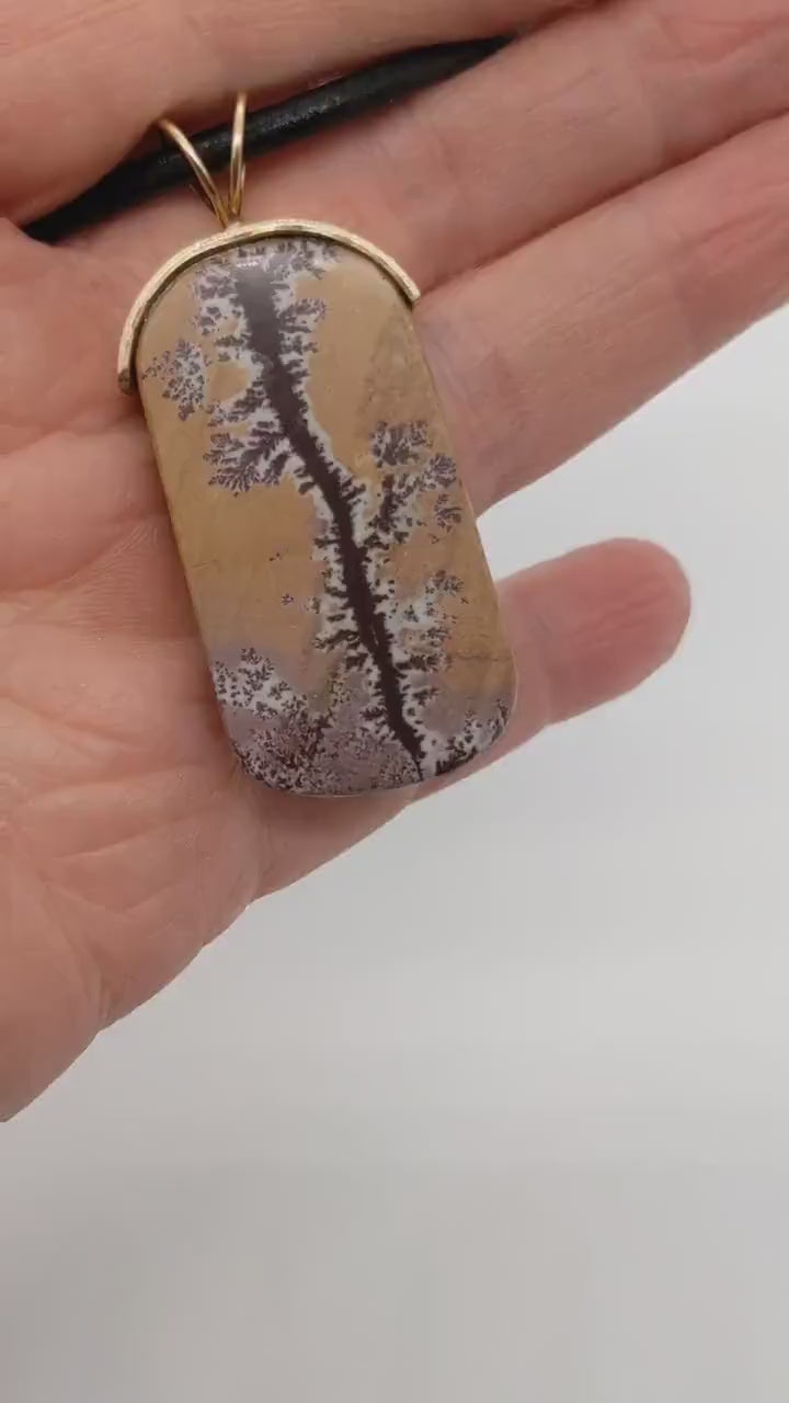 A demo video of a closeup of a one-of-a-kind Sonoran Dendritic Rhyolite Charm Pendant, hand-cut and polished to reveal the natural warm tan and soft pink hues accented by deep brown dendritic branch like patterns.