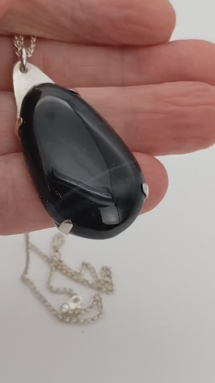 Demo of Handcrafted African Blue Tiger's Eye teardrop pendant with deep blue-black reflective gemstone, solid Argentium 935 silver tabbed setting and silver chain in a hand.