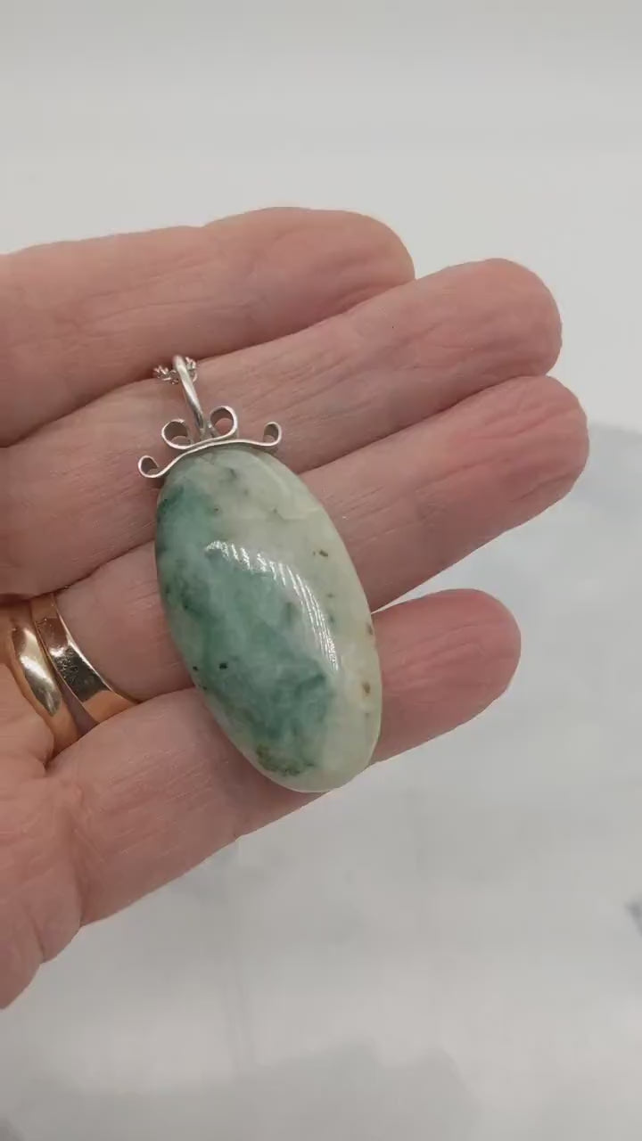 Demo clip in palm of hand to show the features of this African Turquoise Jasper pendant handcut polished on both sides for a reversible design. The stone reveals captivating hues of green and cream, reminiscent of natural turquoise, yet with its own distinct charm.  Set in a handcrafted bow-shaped bail made from tarnish-resistant Argentium® silver