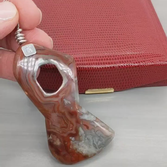 Demo Clip of hand showing a closeup of front of the Crazy Lace Agate features and also the back.  Shown on light gray background with red gift box in background.