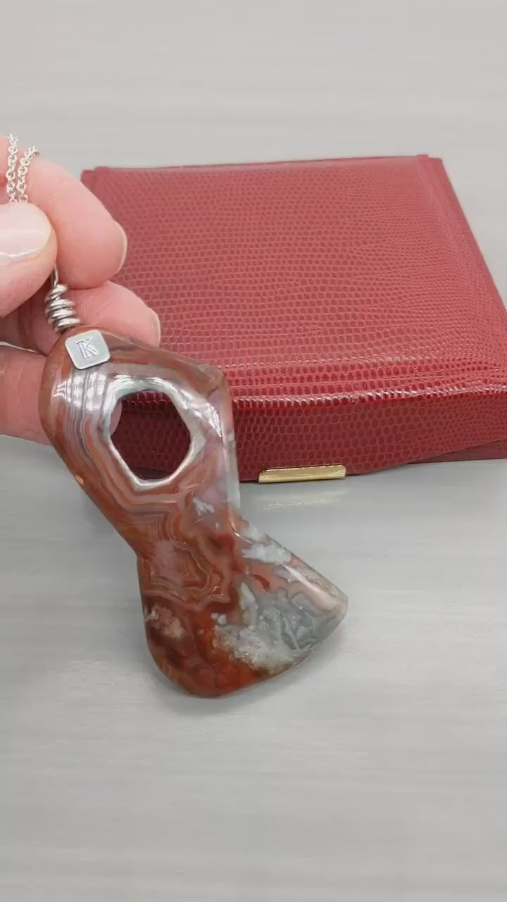 Demo Clip of hand showing a closeup of front of the Crazy Lace Agate features and also the back.  Shown on light gray background with red gift box in background.