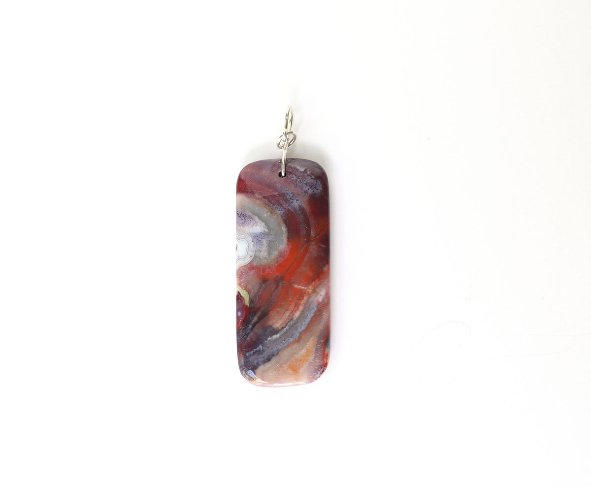 The reverse side of this one-of-a-kind handcut and polished Red Mexico Agate (Natural Banded Agate) Pendant features flowing bands of deep red, purple, orange, black, and white. Shown on white background.