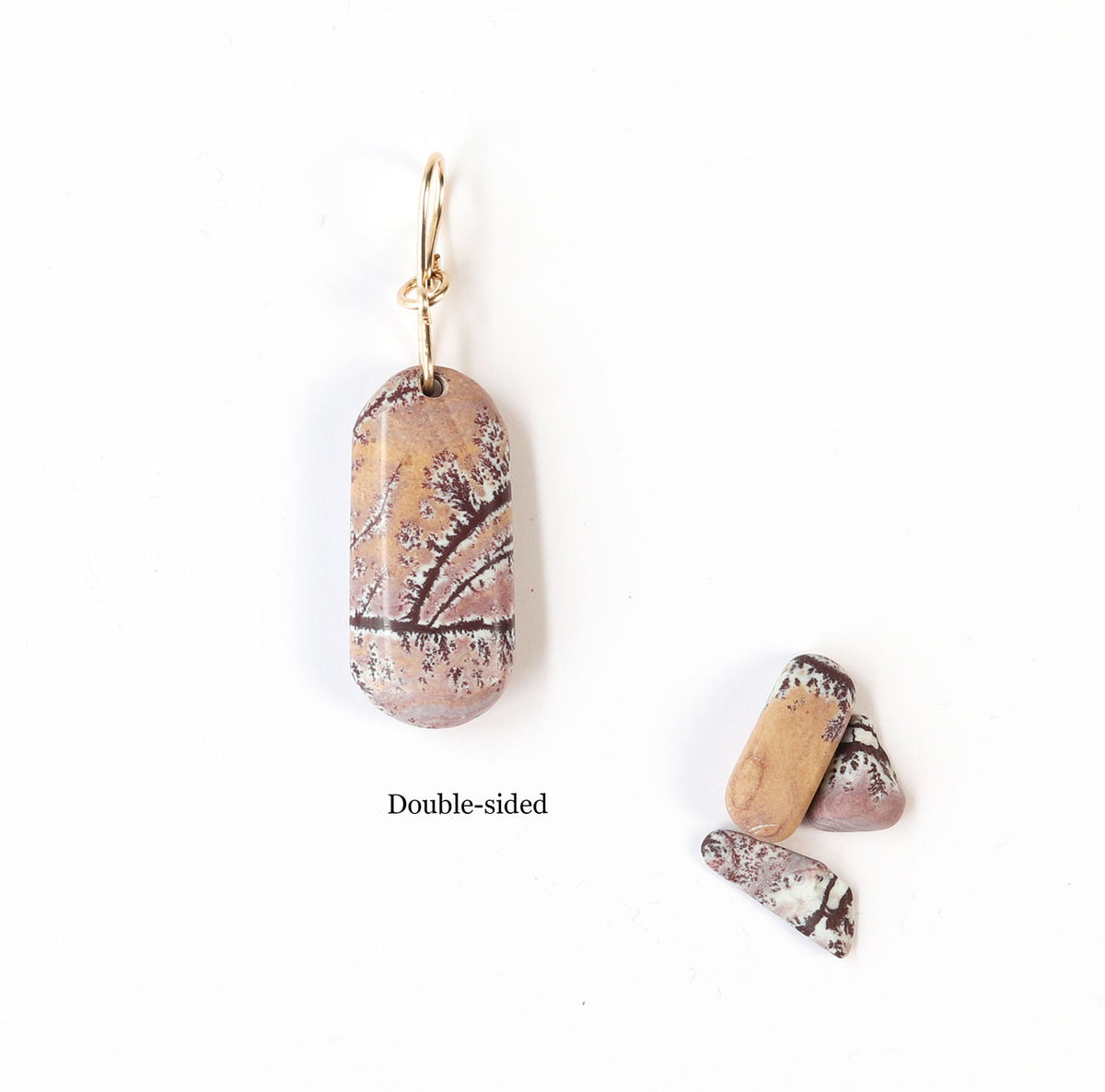 The reverse side is shown of this Sonoran Dendritic Rhyolite pendant charm showcases warm tan and soft pink tones laced with delicate white and deep brown dendritic patterns finished with a custom 14/20 gold-filled infinity bow bail.  Shown on white background next a few raw Sonoran Dendritic pebbles for props.