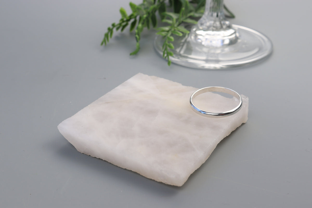 Handcrafted solid Argentium 935 silver half-round ring with smooth polished band displayed beside a clear glass pedestal. displayed on a small white stone stand against a gray background with a glass base and green leaves blurred in the background.