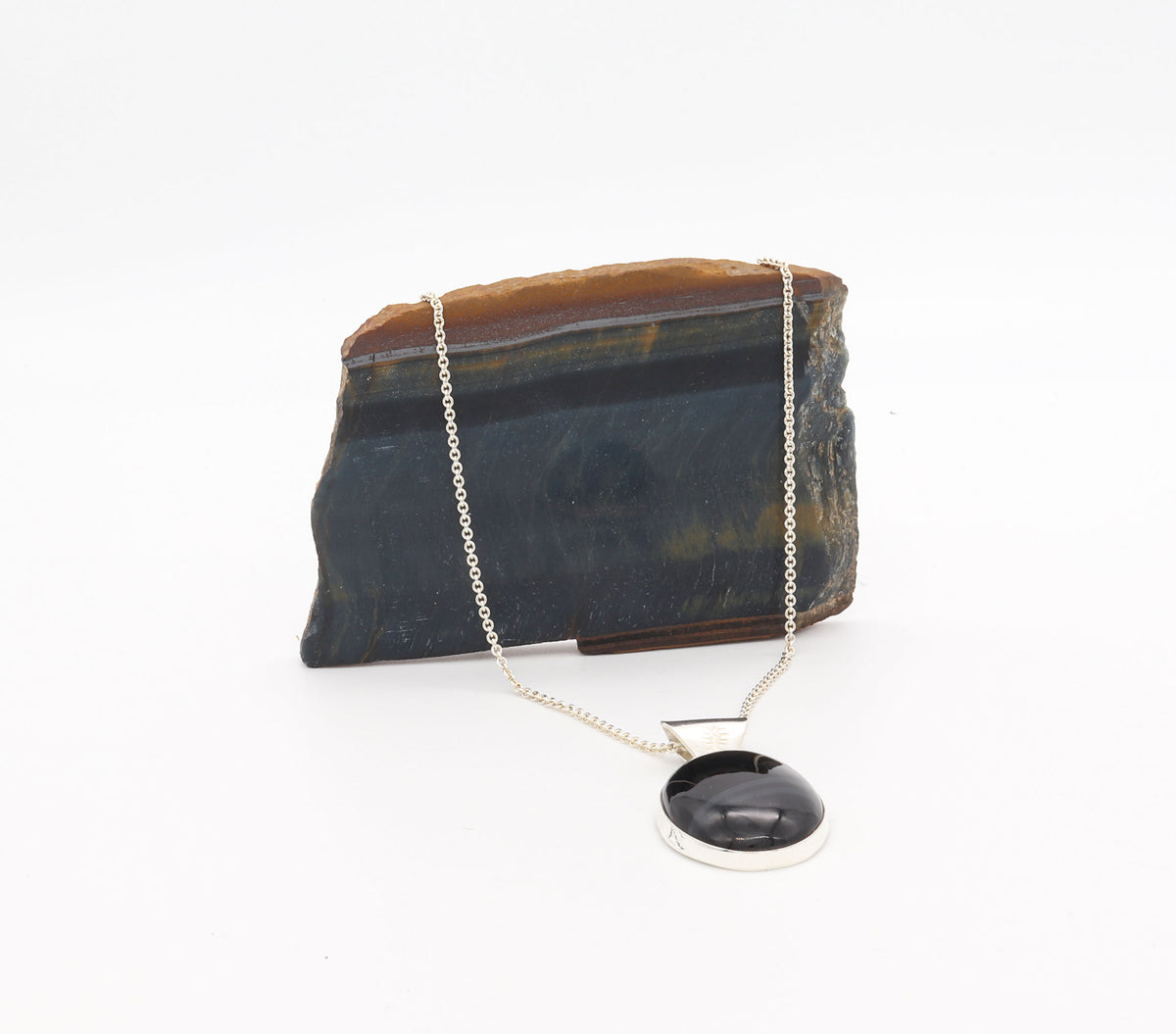 An oval natural Blue Tiger’s Eye pendant in a polished Argentium 935 silver bezel, suspended from a silver chain against a white background, displayed in front of a polished brown and black stone slab on a white background.