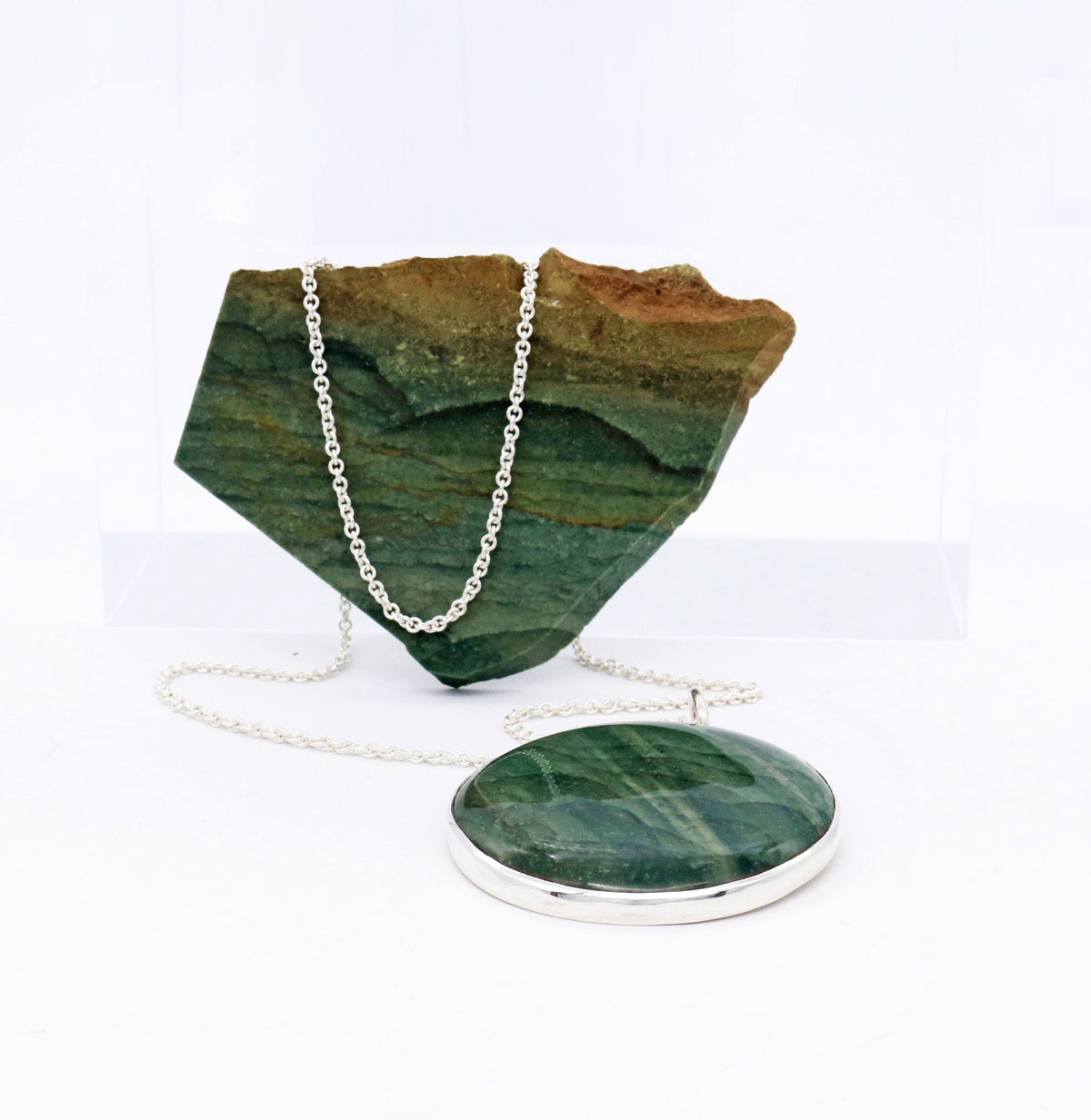 A round green Arizona Lizard stone pendant necklace with a silver bezel and chain, displayed in front of a raw Arizona Lizard green stone display block on a white background.
