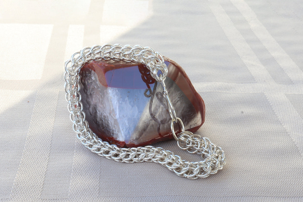 A silver persian weave chain bracelet draped over a polish agate stone with reddish-brown and blue-gray tones, resting on a white textured fabric surface.