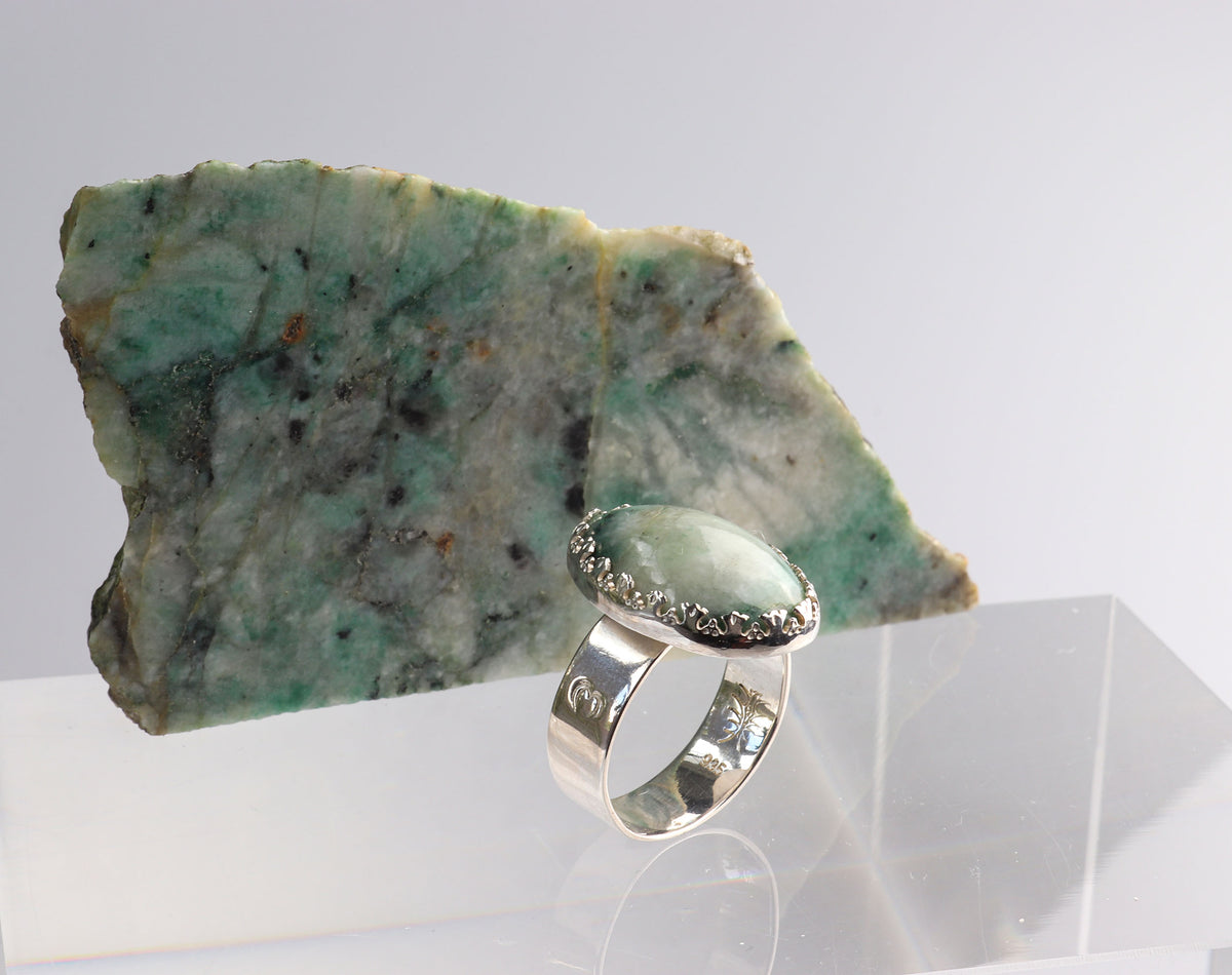 African Turquoise Jasper ring handcrafted in polished Argentium 935 silver, displayed on a clear acrylic block in front of a rough African Turquoise Jasper rock against a light gray background.