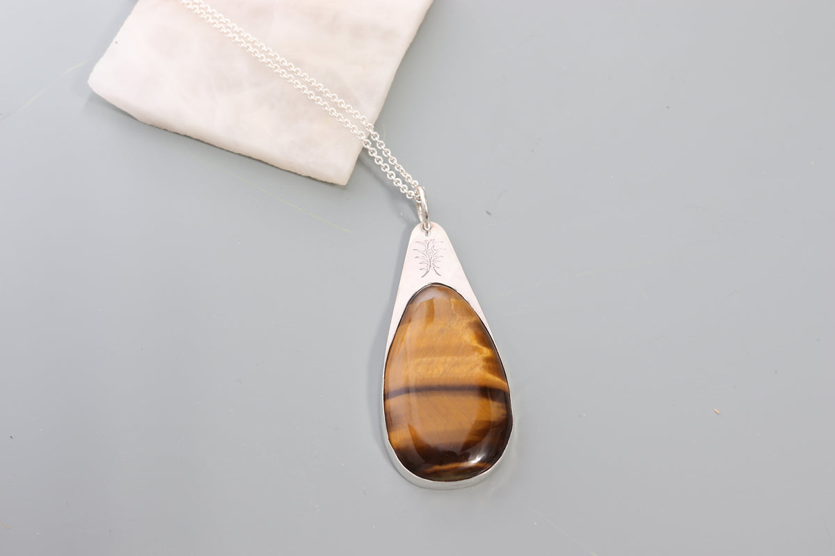 A handcrafted Tiger's Eye Pendant polished to bring out its unique silky wavy brilliance and natural chatoyancy. Set in a custom Argentium® .935 Silver bezel and paired with a complementary silver 935 Argentium necklace chain shown next to white rock on a light gray background.
