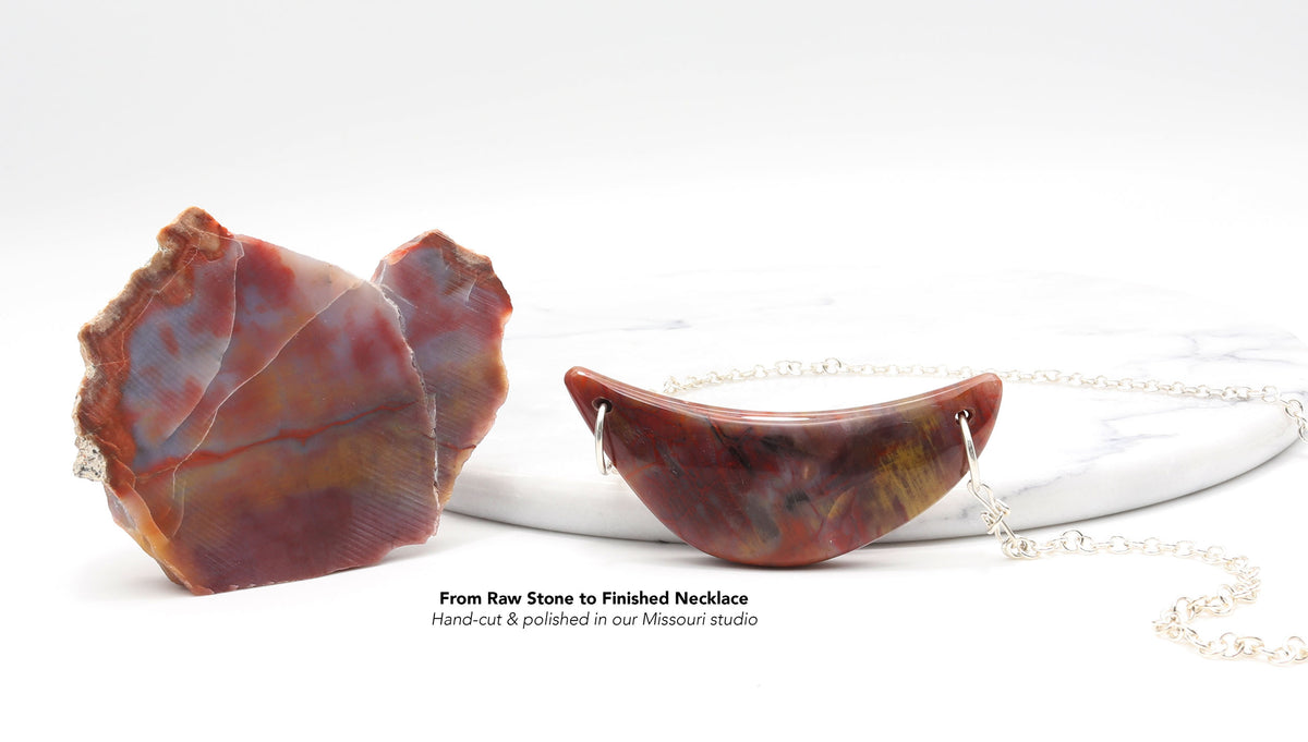 A polished crescent-shaped raw Petrified Wood stone pendant necklace in reddish-brown and amber tones, shown next to its rougher raw stone form on a white marble slab with a silver chain. Text overlay reads 'From Raw Stone to Finished Necklace Hand-cut & polished in our Mission studio'.