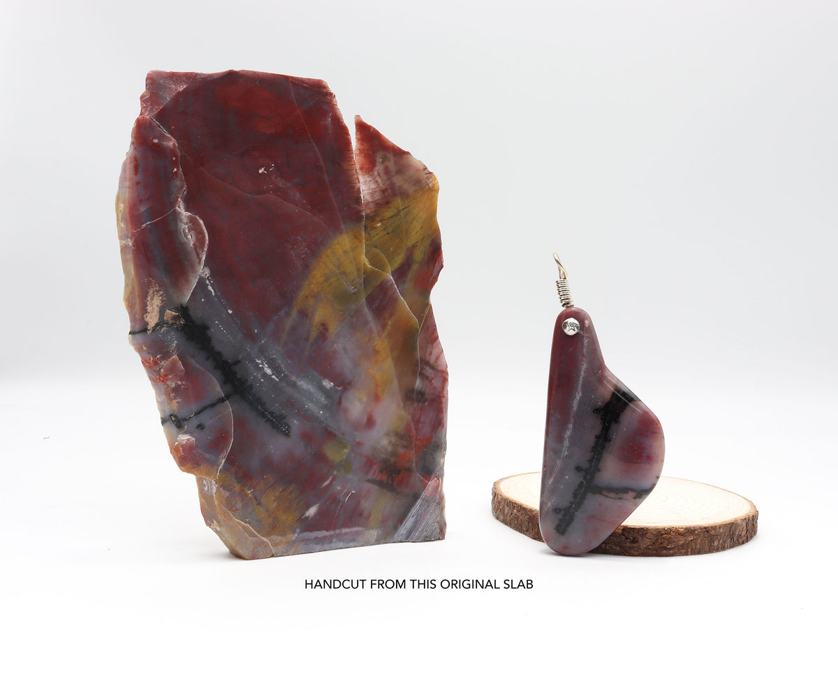 Handcrafted Arizona Petrified Rainbow Wood charm, 4 inches long, with a 935 silver bail, on a white background, next to the raw Petrified Rainbow Wood rock used to craft it, showcasing the transformation from raw rock to a one-of-a-kind gemstone with bold red and carbon black stripes and natural swirls.  Shown on white background. Pendant is shown leaning on a slab of wood as a prop.