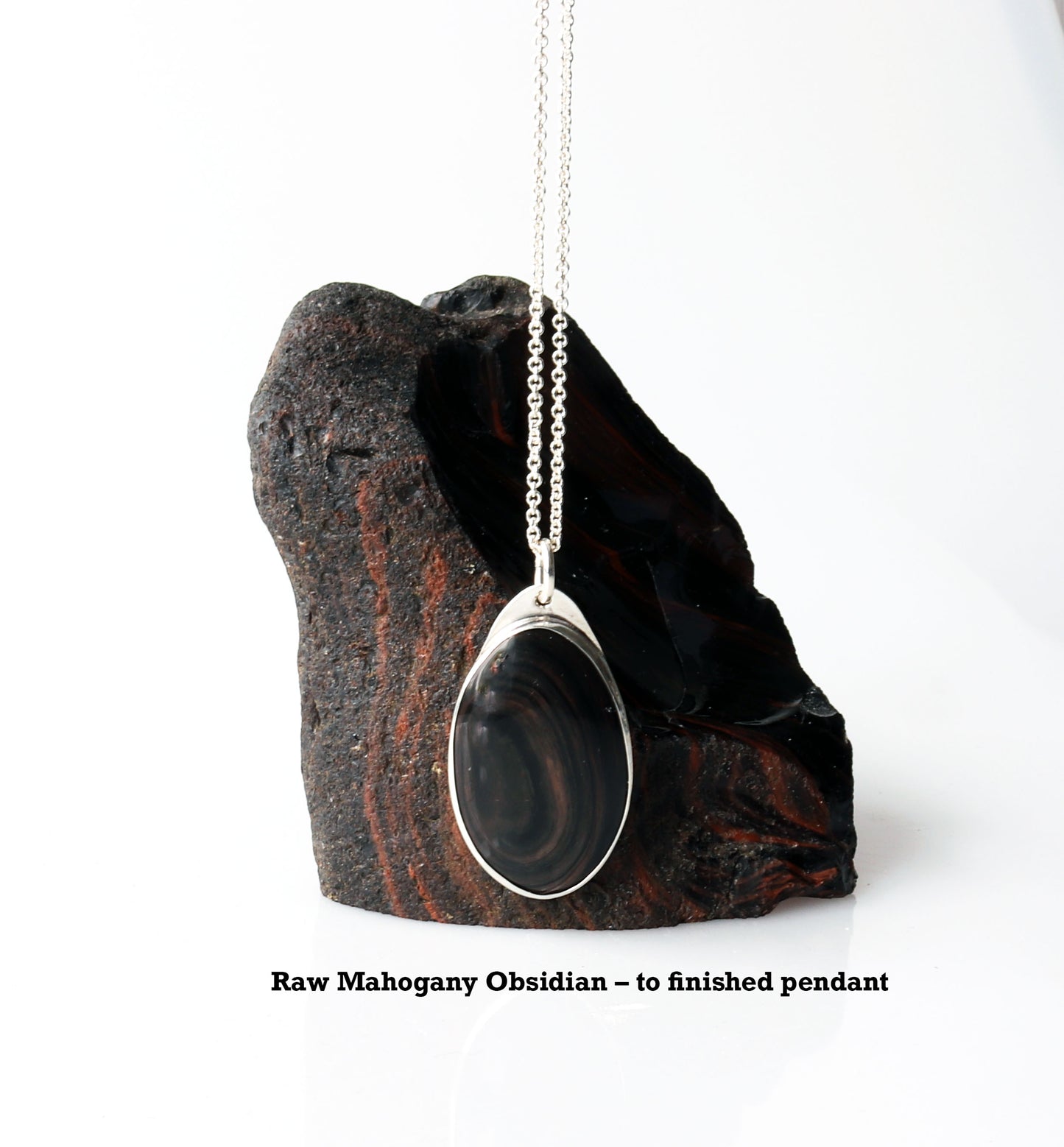 A Mahogany Obsidian pendant with swirling patterns in deep reddish-brown and dark mahogany hues,  set in Argentium® 935 Silver.  Shown next to raw Mahogany Obsidian rock used to craft this pendant.