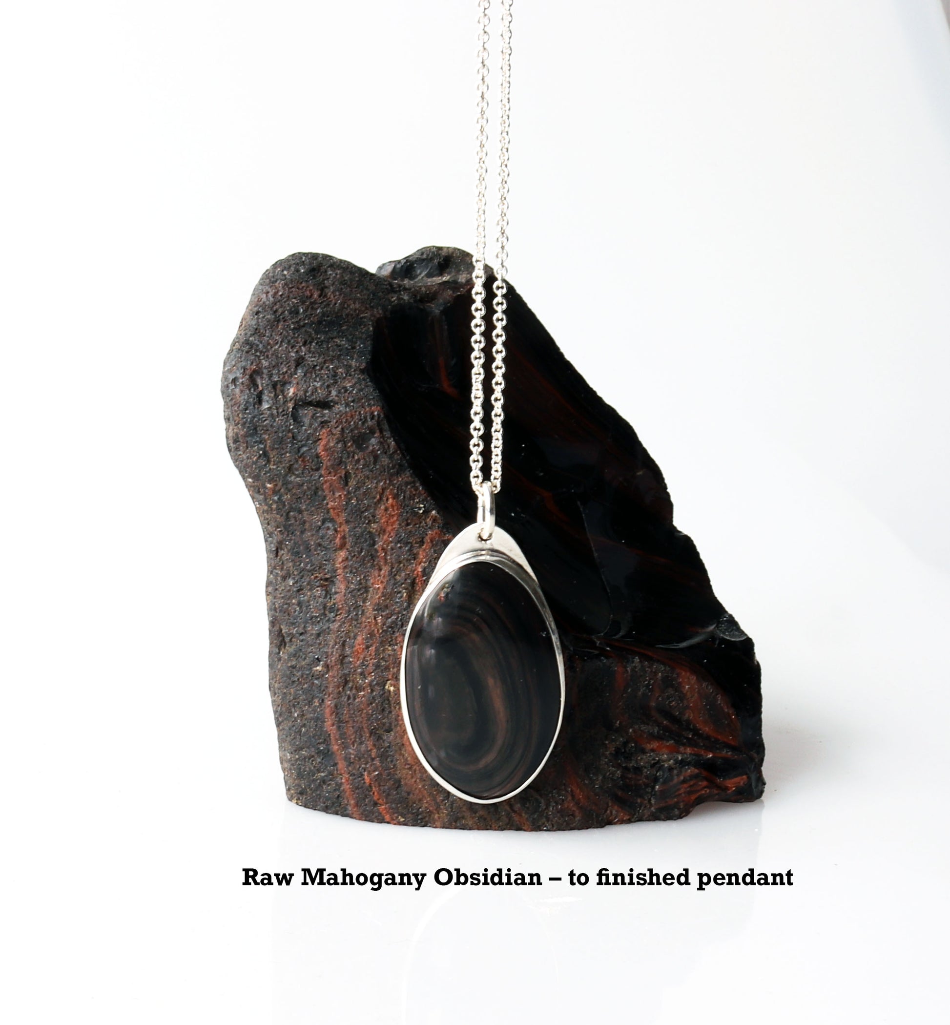 A Mahogany Obsidian pendant with swirling patterns in deep reddish-brown and dark mahogany hues,  set in Argentium® 935 Silver.  Shown next to raw Mahogany Obsidian rock used to craft this pendant.