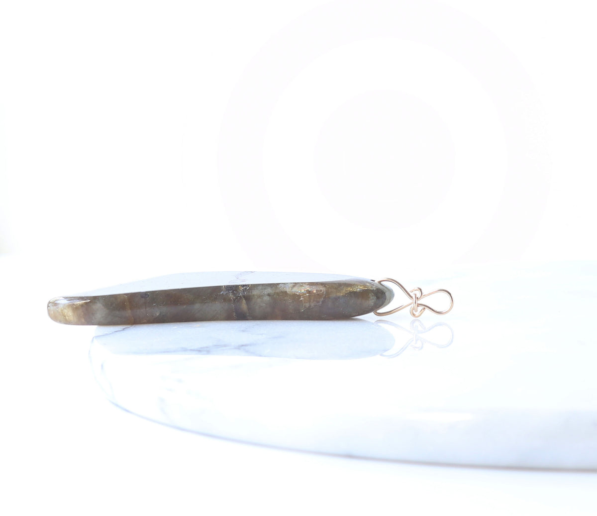 A flat, elongated stone pendant in earthy brown and gray tones with a delicate gold wire loop, resting on a white marble slab against a bright white background.