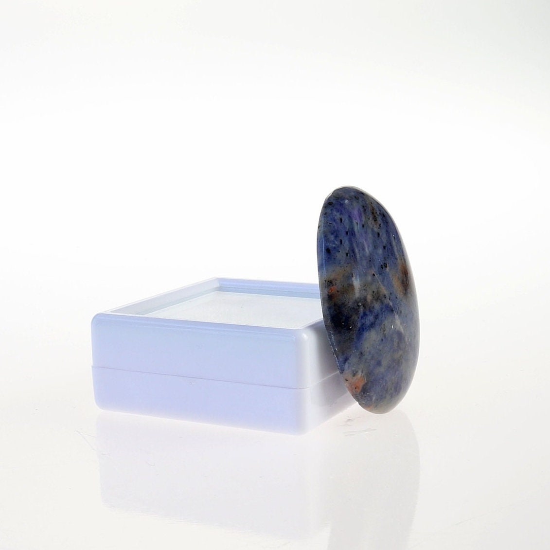 A side view of this hand-cut Blue Foidolite Sodalite oval cabochon with mottled blue, creamy white, gray, and warm brown natural patterns.  Shown against a white gem box against a white background.