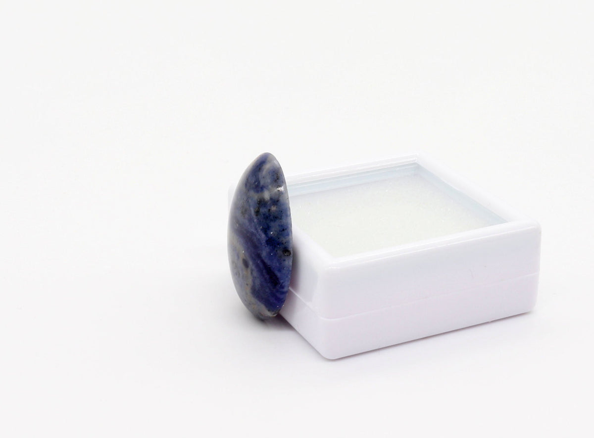 A hand-cut and polished natural Foidolite cabochon with layered deep blue, slate blue and gray natural mineral patterning displayed at side angle against a white gem box, photographed on a clean white background.