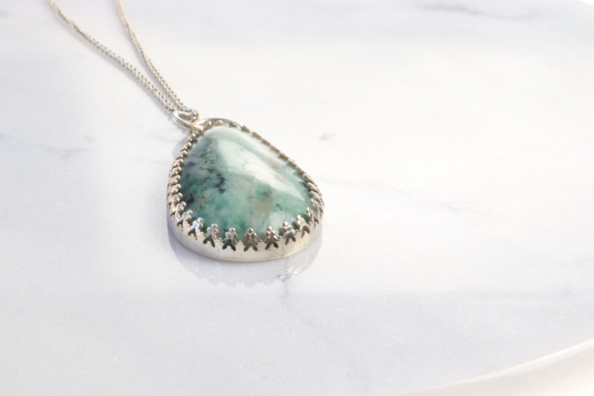 A closeup of this green marbled natural African Turquoise Jasper pendant and the decorative gallery-edge setting that frames the stone while resting on a white marble surface.