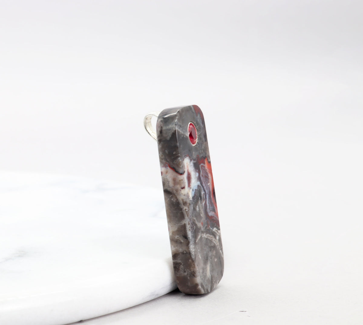 A side view of Red Mexican Agate pendant with 6mm lab-grown ruby inlay and Argentium Silver bail shown against a white marble slab on white background.