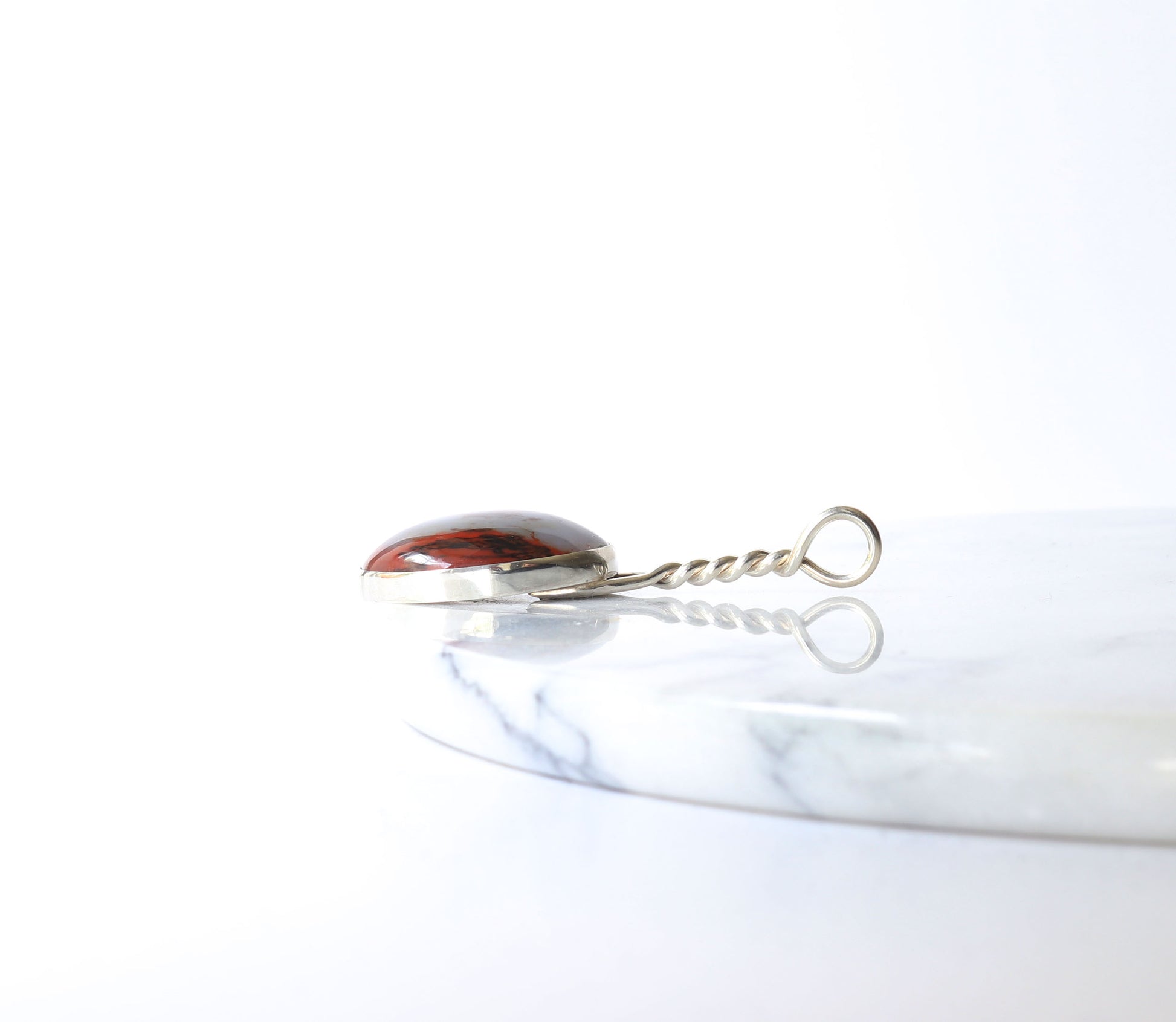  A side view of a natural petrified rainbow wood pendant  set in solid Argentium® Silver with a handcrafted twist bail shown on white marble against light gray background.