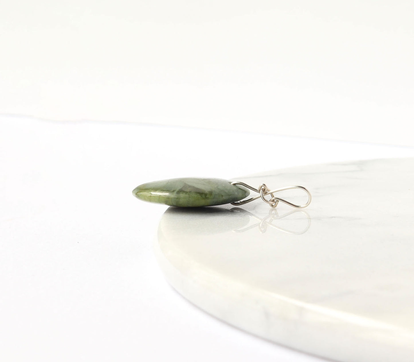 A polished green stone pendant with a silver wire-wrapped hook, resting on a white marble slab against a white background.