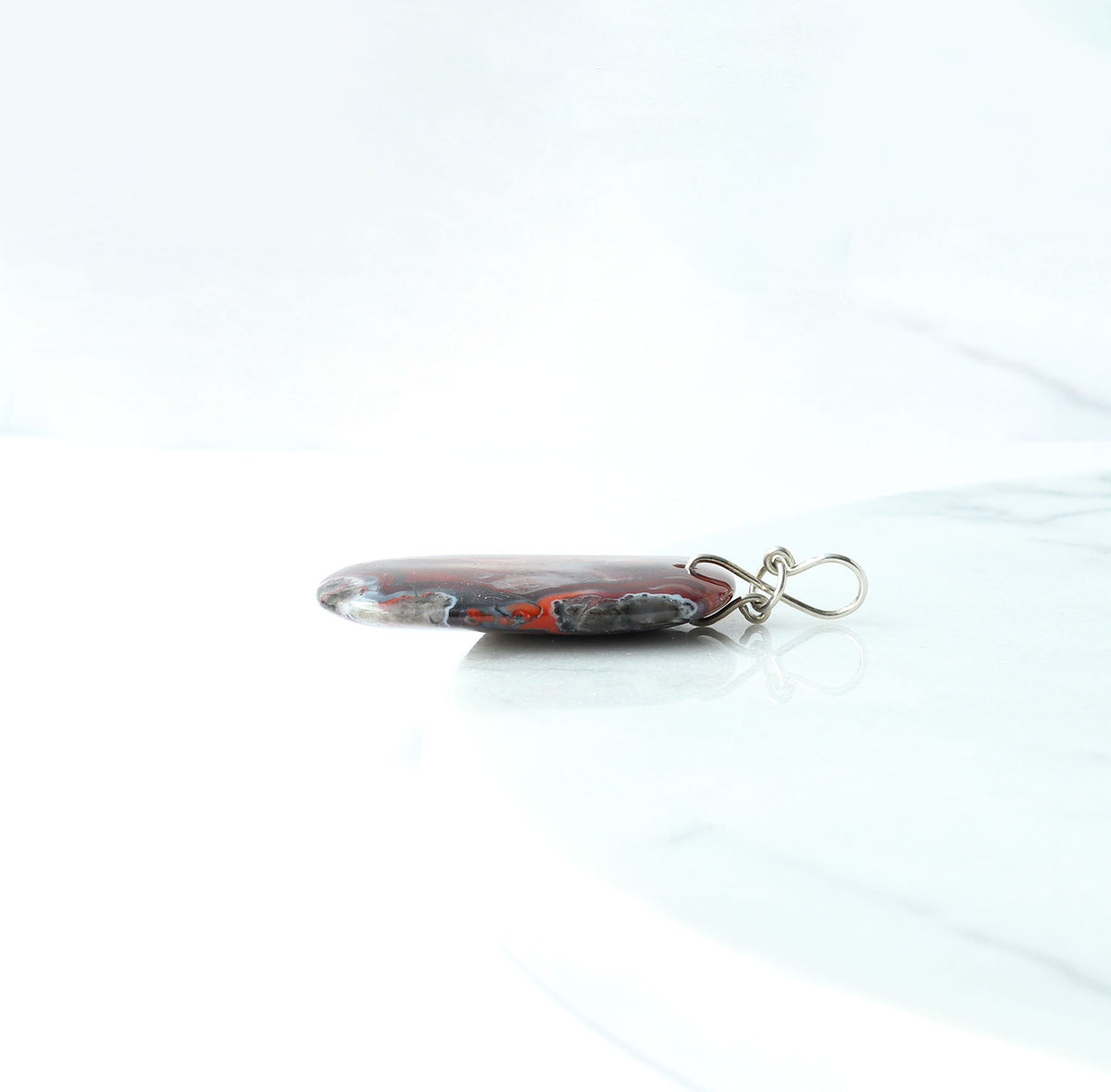 Side view of this Red Mexican Agate pendant, hand-cut and polished on both sides with custom handcrafted infinity bow bail in Argentium silver bail suspended from optional 1.9 oval Argentium chain.  Shown on white background on a white marble slab.