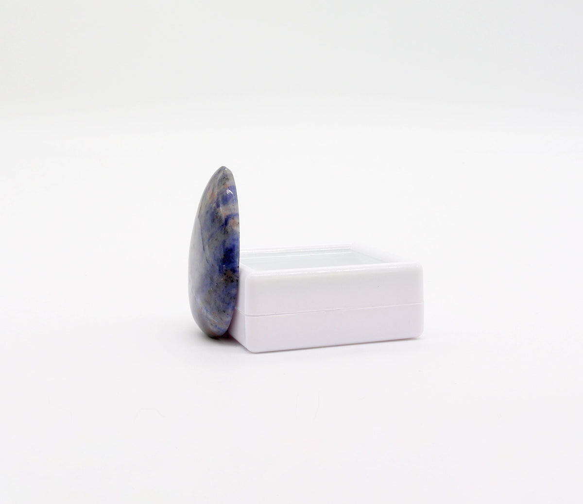 A side-view of a polished teardrop-shaped  Blue Foidolite cabochon, hand-cut and polished in our Missouri studio to showcase its natural blue, gray, cream, and subtle rust-colored patterning.  Shown against a white gem box against white background against a seamless white background.