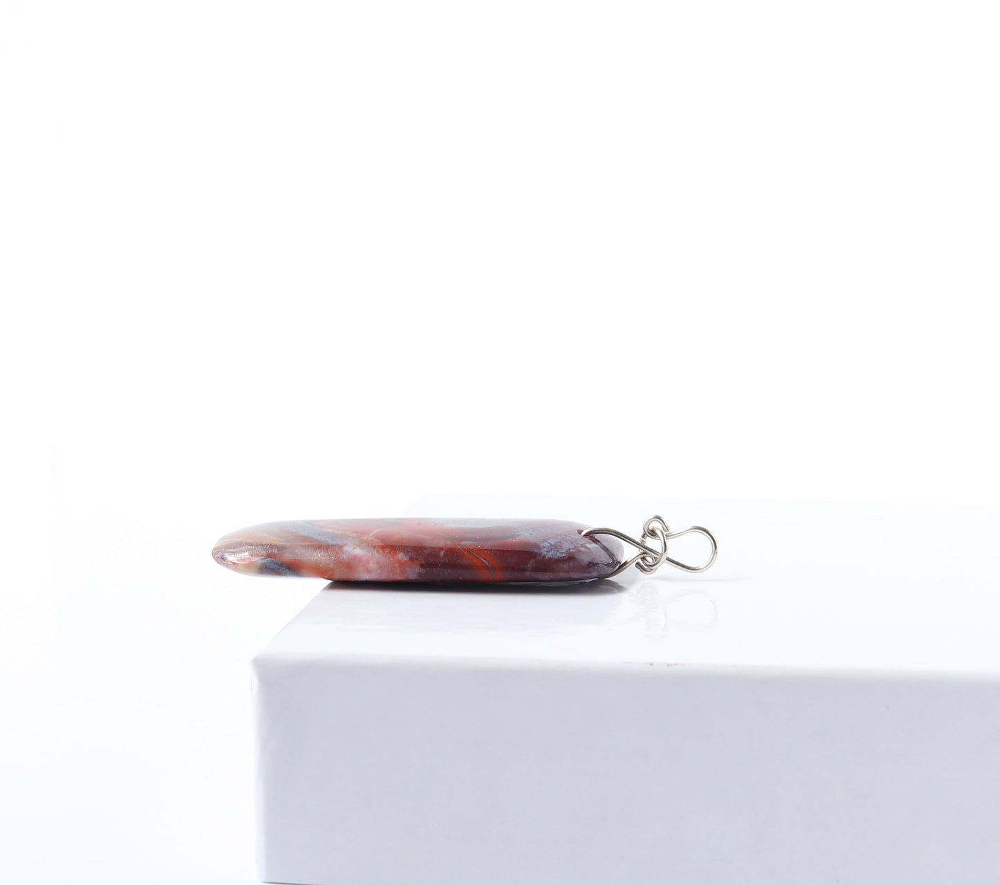 The side view to highlight the infinity bow bail shape in Argentium silver.  This one-of-a-kind handcut and polished Red Mexico Agate (Natural Banded Agate) Pendant features flowing bands of deep red, purple, orange, black, and white. Shown on white background.