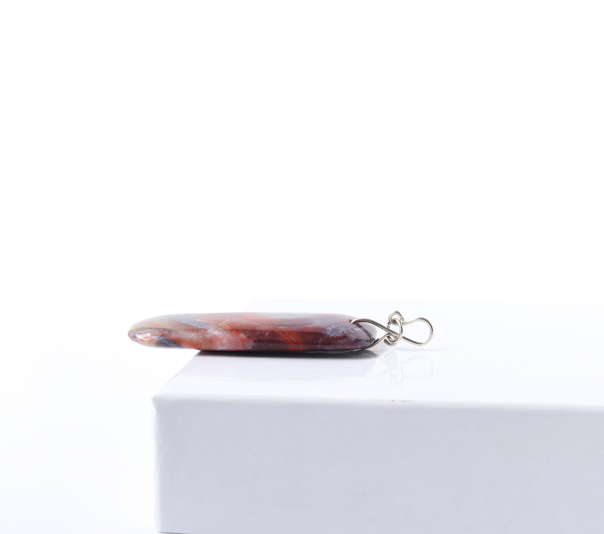 The side view to highlight the infinity bow bail shape in Argentium silver.  This one-of-a-kind handcut and polished Red Mexico Agate (Natural Banded Agate) Pendant features flowing bands of deep red, purple, orange, black, and white. Shown on white background.