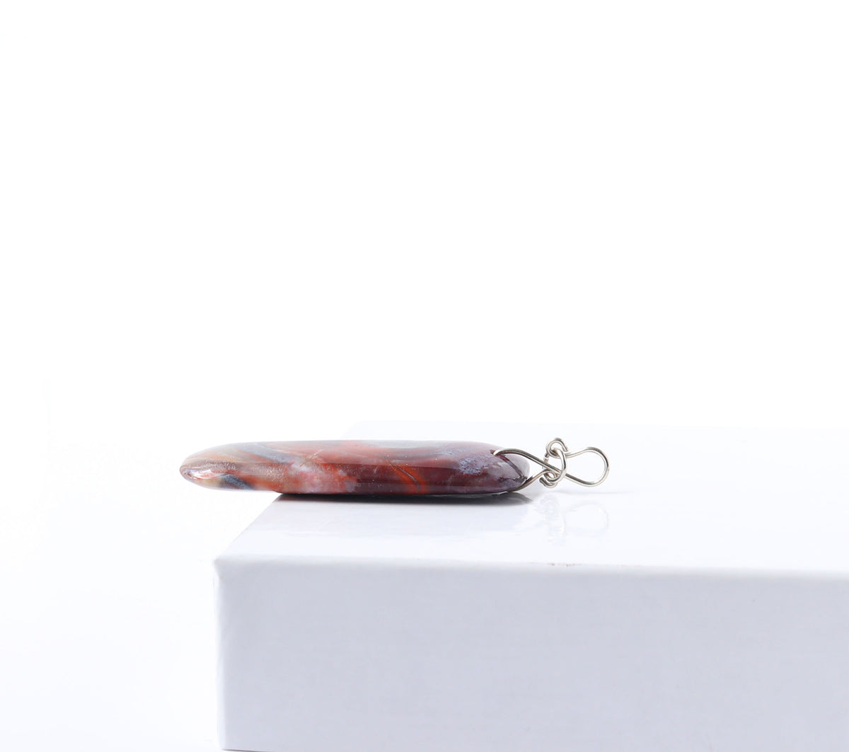 The side view to highlight the infinity bow bail shape in Argentium silver.  This one-of-a-kind handcut and polished Red Mexico Agate (Natural Banded Agate) Pendant features flowing bands of deep red, purple, orange, black, and white. Shown on white background.