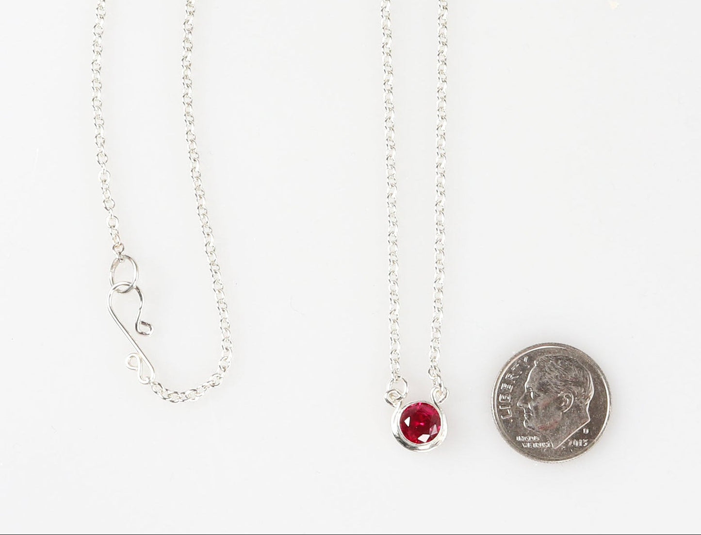 6mm round faceted lab-grown ruby nestled in a custom Argentium® Silver bezel setting.  Shown next U.S. dime for scale against white background.