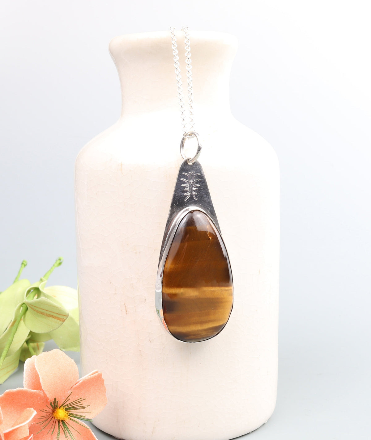 A handcrafted Tiger's Eye Pendant polished to bring out its unique silky wavy brilliance and natural chatoyancy. Set in a custom Argentium® .935 Silver bezel and paired with a complementary silver 935 Argentium necklace chain hanging from a white ceramic vase with decorative peach flowers on a light gray background.